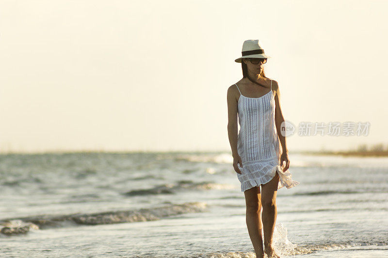 美丽宁静的女人漫步在海滩上，穿着白色连衣裙，草帽和太阳镜在São米格尔dos Milagres，阿拉格阿斯，巴西